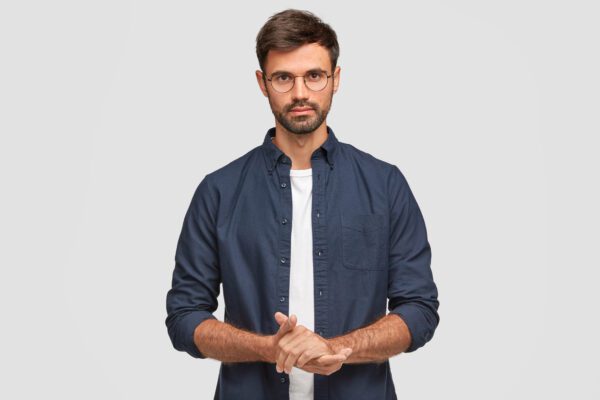 Waist up portrait of handsome serious unshaven male keeps hands together, dressed in dark xanh lơ shirt, has talk with interlocutor, stands against white background. Self confident man freelancer